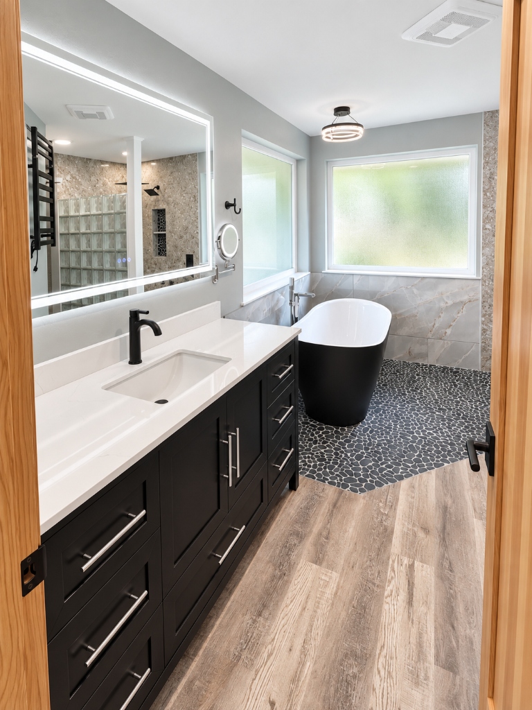 Remodeled bathroom with black shaker vanity, backlit LED mirror, freestanding black soaking tub and pebble tile floor in Kitsap County, WA