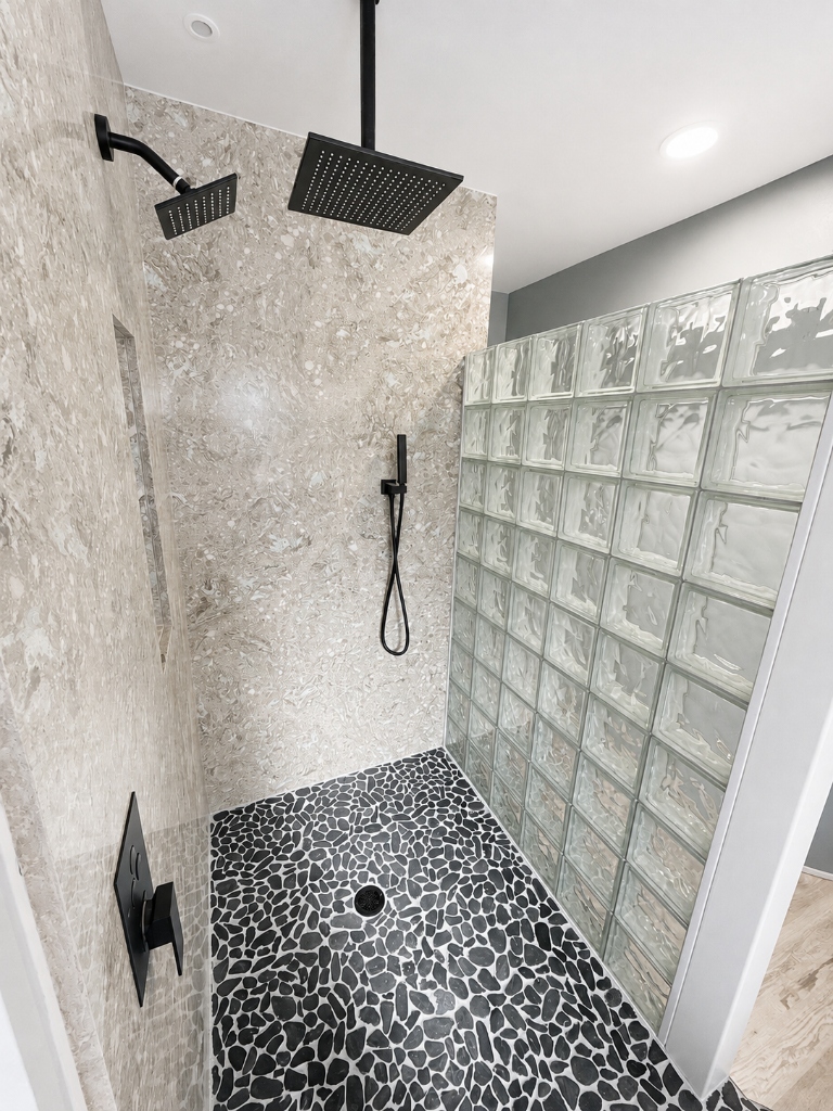 Custom walk-in shower with dual matte black rain shower heads, stone-look wall panels, glass block privacy wall and black pebble tile floor in Kitsap County, WA